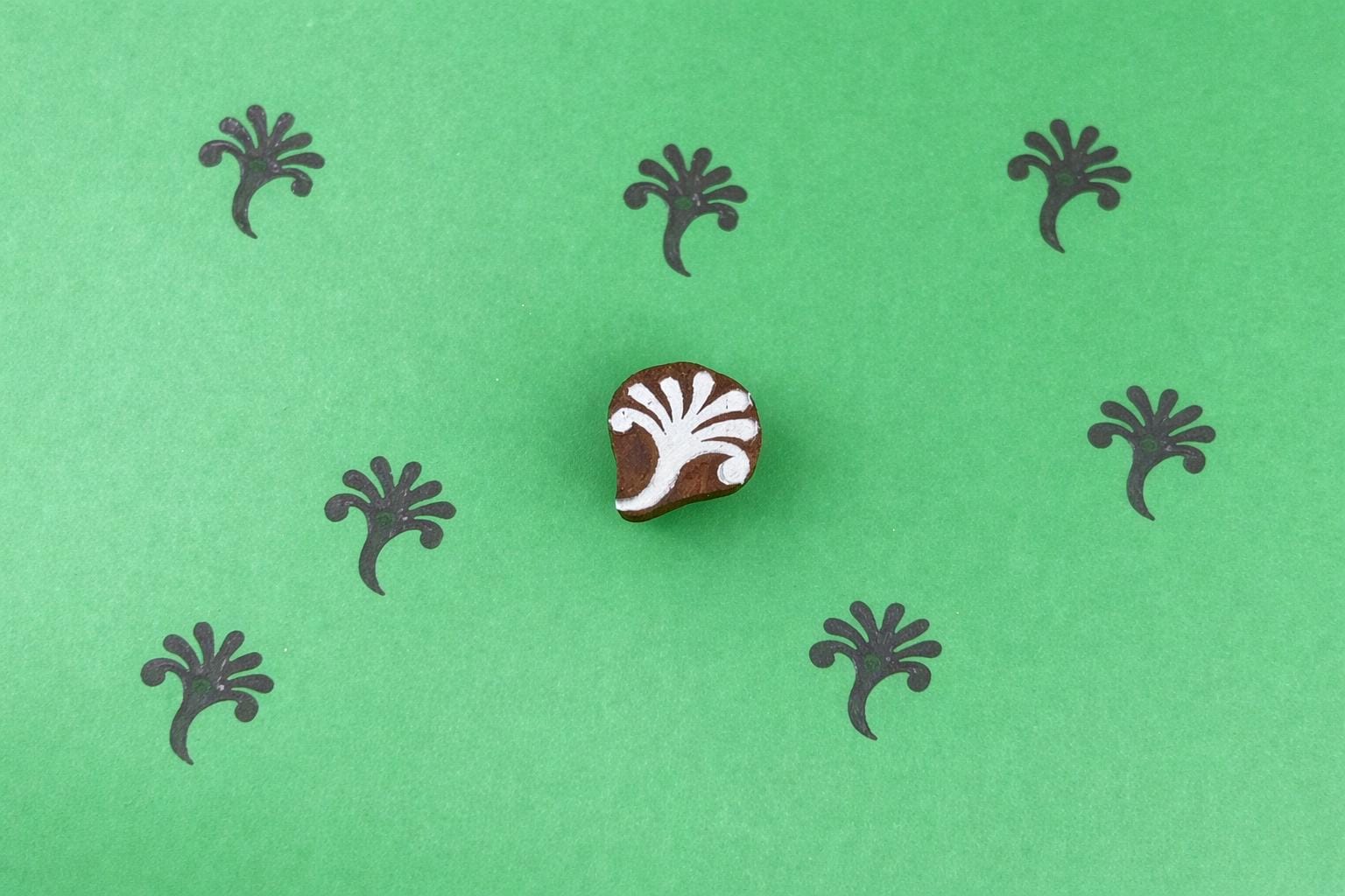 Small hand carved wooden block stamp with abstract botanical sprig design printed in repeating pattern on green background