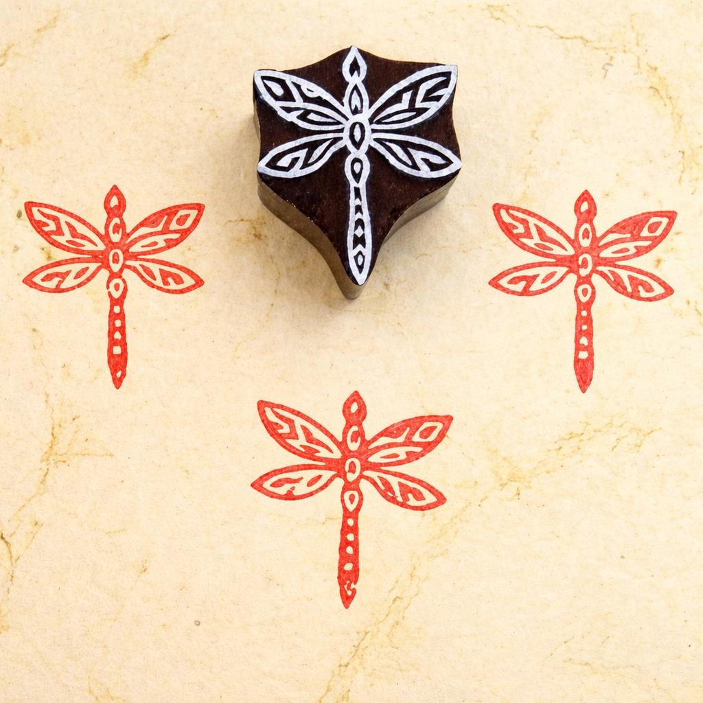 Hand carved wooden block stamp with geometric dragonfly design shown with red printed impressions on textured paper