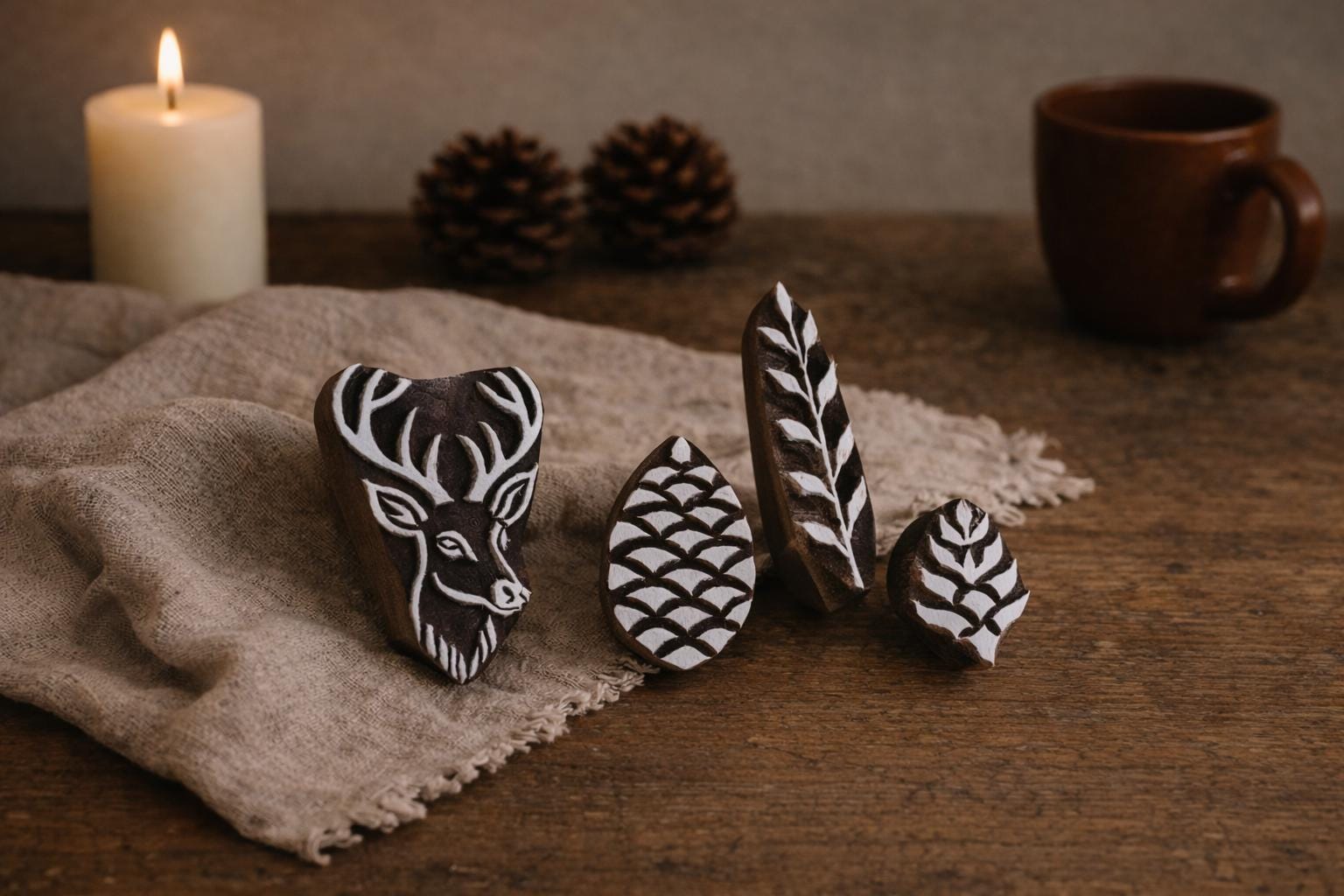 Hand carved wooden stamp set with deer head, pinecone and botanical leaf designs arranged on rustic fabric with candle and pinecones