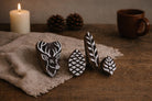 Hand carved wooden stamp set with deer head, pinecone and botanical leaf designs arranged on rustic fabric with candle and pinecones