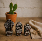 Hand carved cactus stamp set with three designs on rustic wooden surface with potted cactus in background