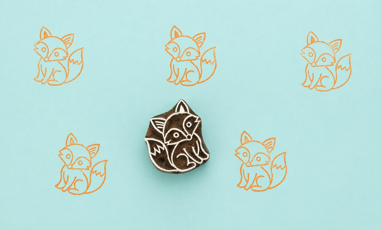 Hand carved wooden fox printing stamp with repeated orange fox impressions on teal background, cute woodland animal design for fabric and paper printing