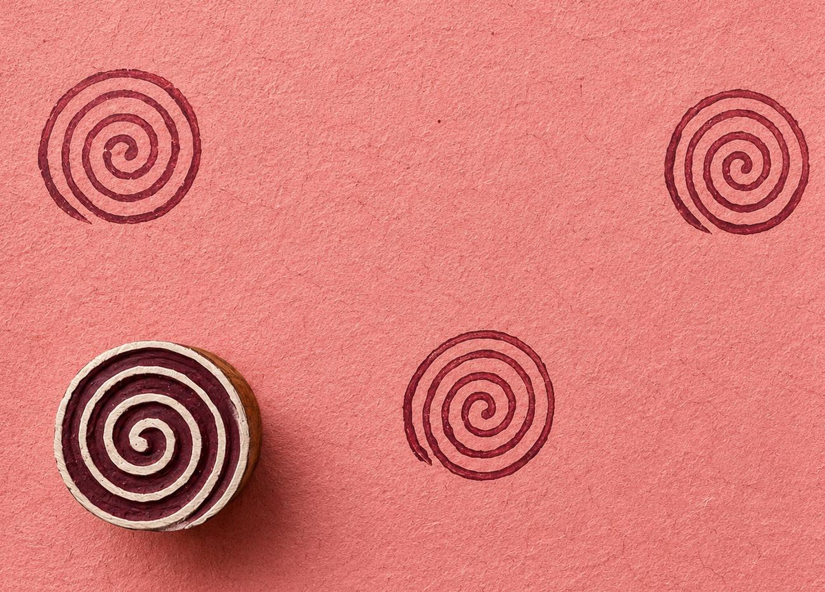 Hand carved wooden spiral block stamp printing repeating swirl motif in burgundy ink on handmade pink paper
