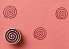 Hand carved wooden spiral block stamp printing repeating swirl motif in burgundy ink on handmade pink paper