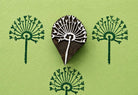 Hand carved wooden printing block featuring a radiating botanical meadow burst motif stamped repeatedly in dark green ink on textured green paper.