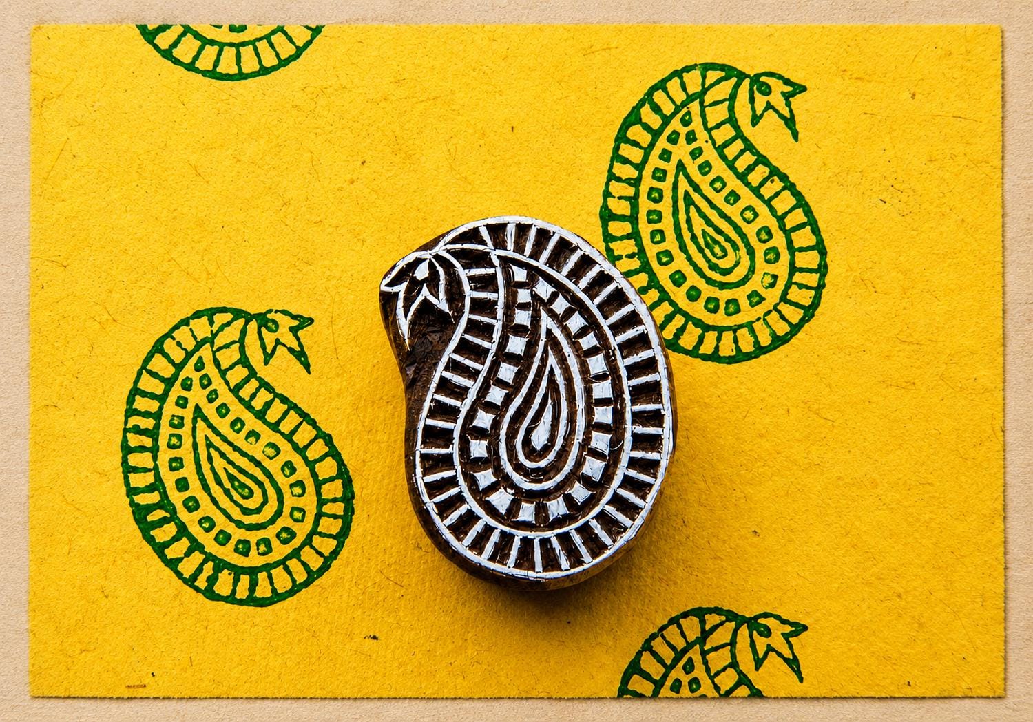 Hand carved wooden paisley block stamp printing repeating green paisley motif on textured yellow handmade paper