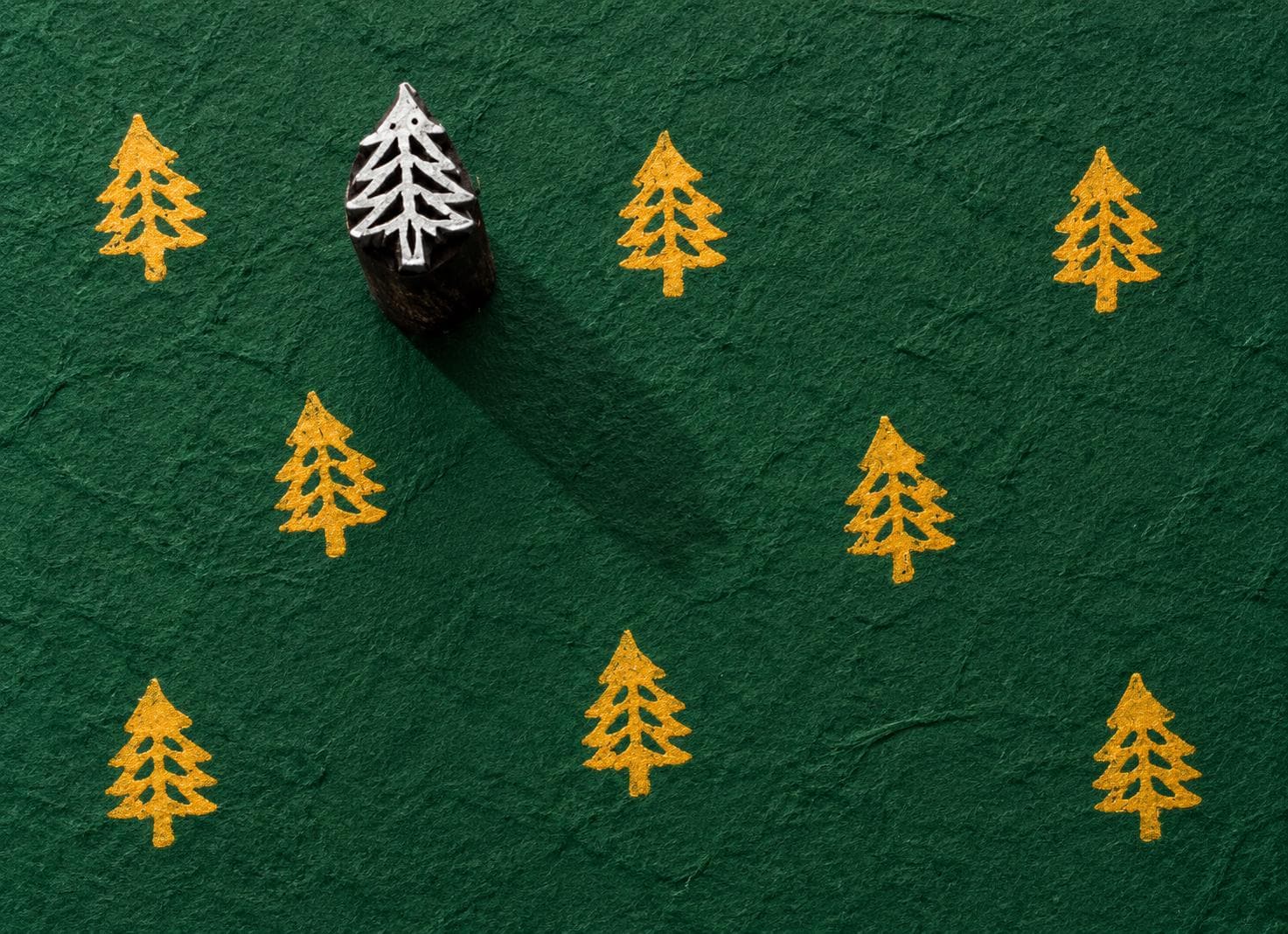 Tiny hand carved wooden evergreen tree block stamp printing repeating pine tree motif in golden yellow ink on textured forest green paper