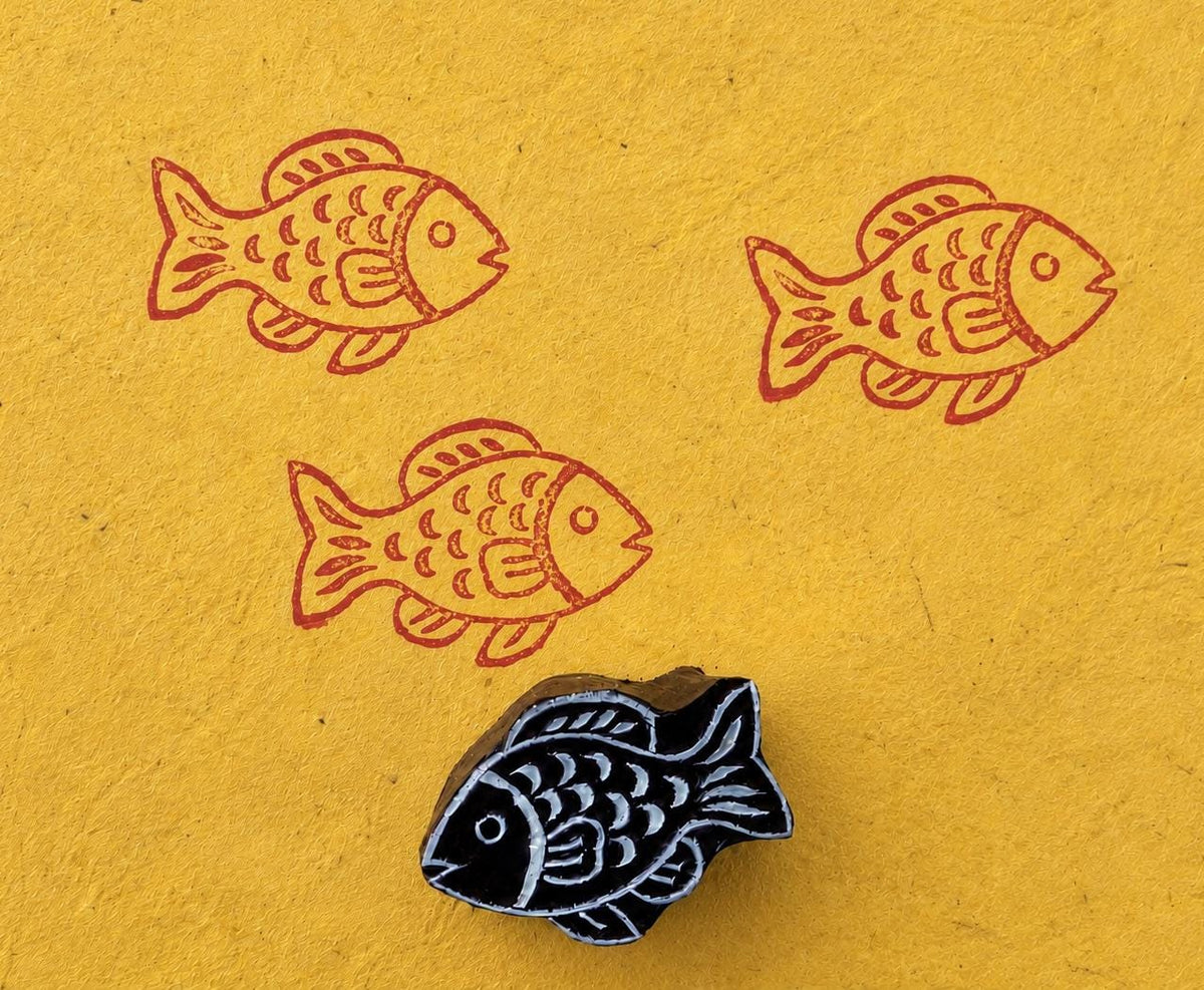 Hand carved wooden fish block stamp printing repeating tropical fish motif in coral red ink on textured yellow paper