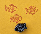 Hand carved wooden fish block stamp printing repeating tropical fish motif in coral red ink on textured yellow paper
