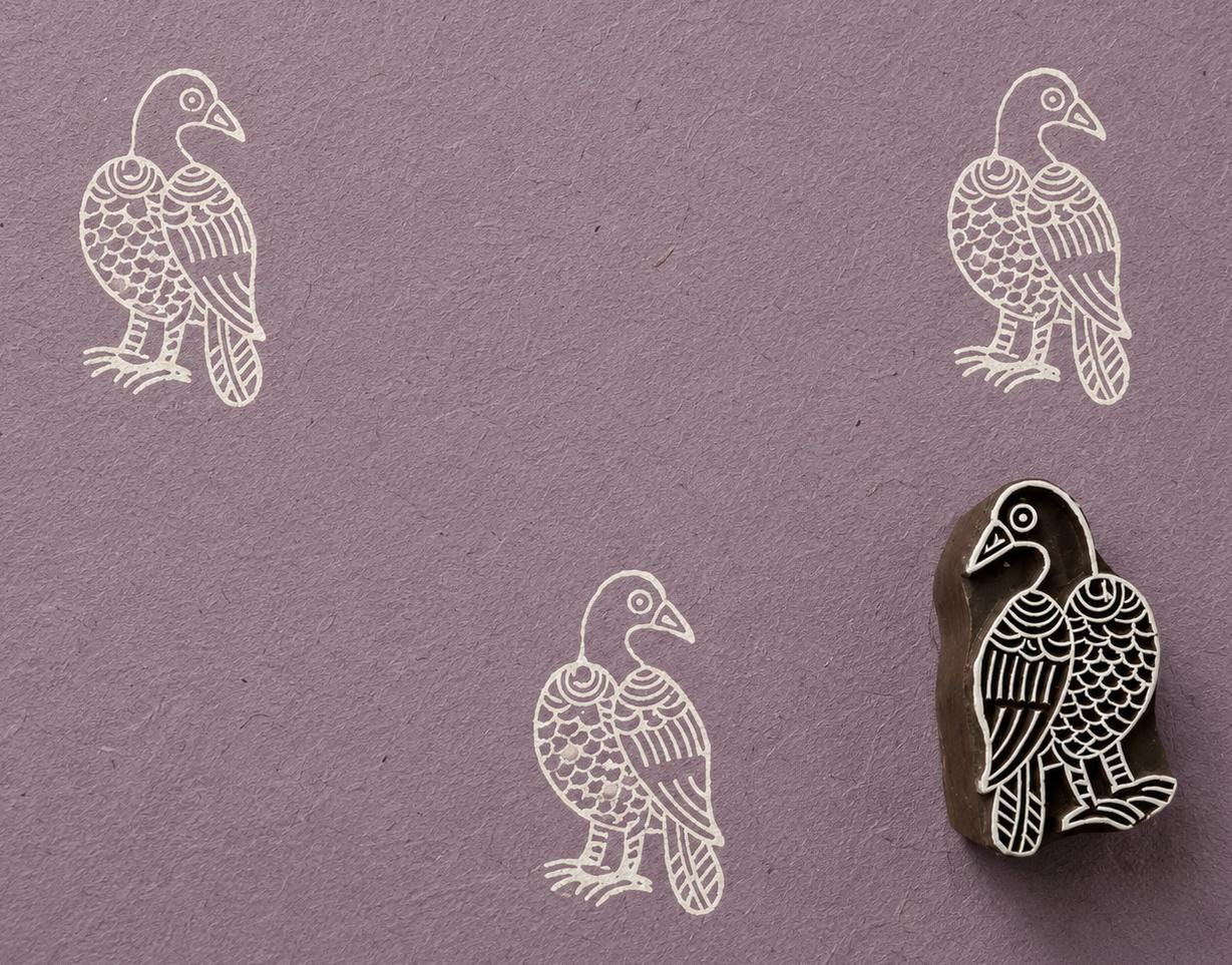 Hand-carved wooden printing block stamp featuring a folk-art raven design, shown with ivory bird impressions on textured lavender handmade paper.