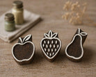 Set of three hand carved wooden fruit printing blocks featuring apple, strawberry, and pear designs arranged on rustic linen fabric background for textile printing and craft projects.
