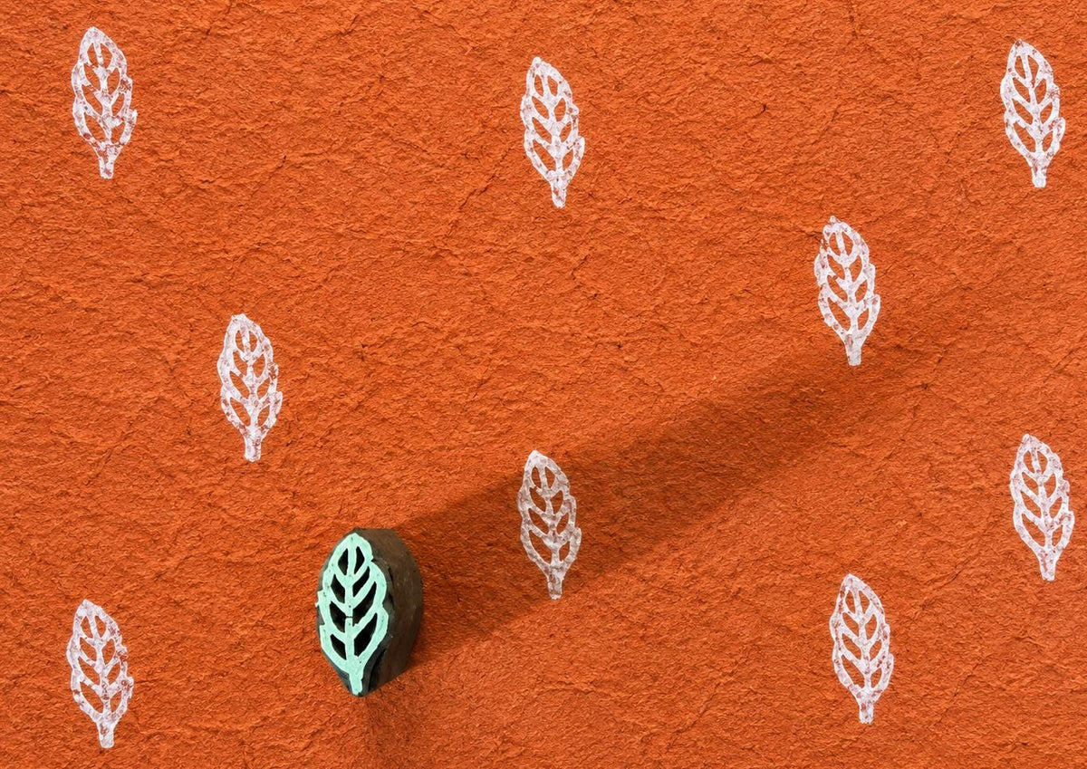 Tiny hand carved wooden leaf sprig block stamp printing delicate repeating botanical motif in white ink on textured terracotta paper