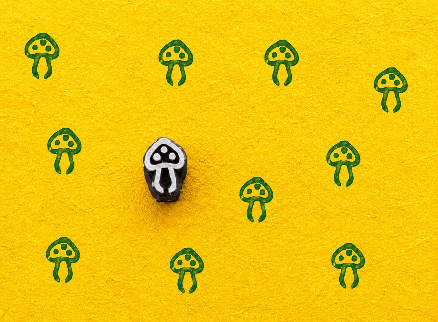 Tiny hand carved wooden block stamp featuring a whimsical toadstool mushroom motif printed repeatedly on textured yellow handmade paper.