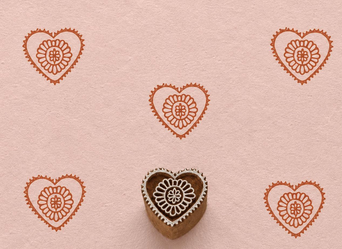 Hand-carved wooden printing block featuring a heart-shaped motif with a floral rosette center stamped in terracotta ink on blush handmade paper.