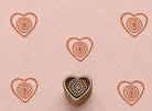 Hand-carved wooden printing block featuring a heart-shaped motif with a floral rosette center stamped in terracotta ink on blush handmade paper.