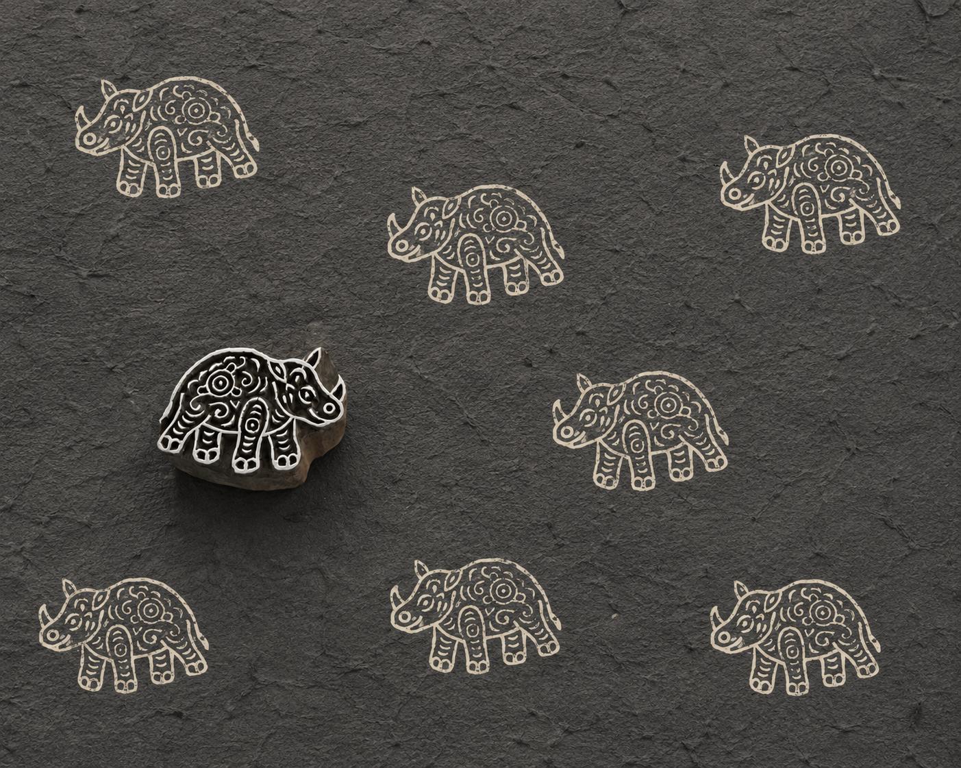 Hand-carved wooden printing block featuring a folk art rhinoceros design stamped repeatedly in ivory ink on textured charcoal handmade paper.