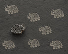 Hand-carved wooden printing block featuring a folk art rhinoceros design stamped repeatedly in ivory ink on textured charcoal handmade paper.