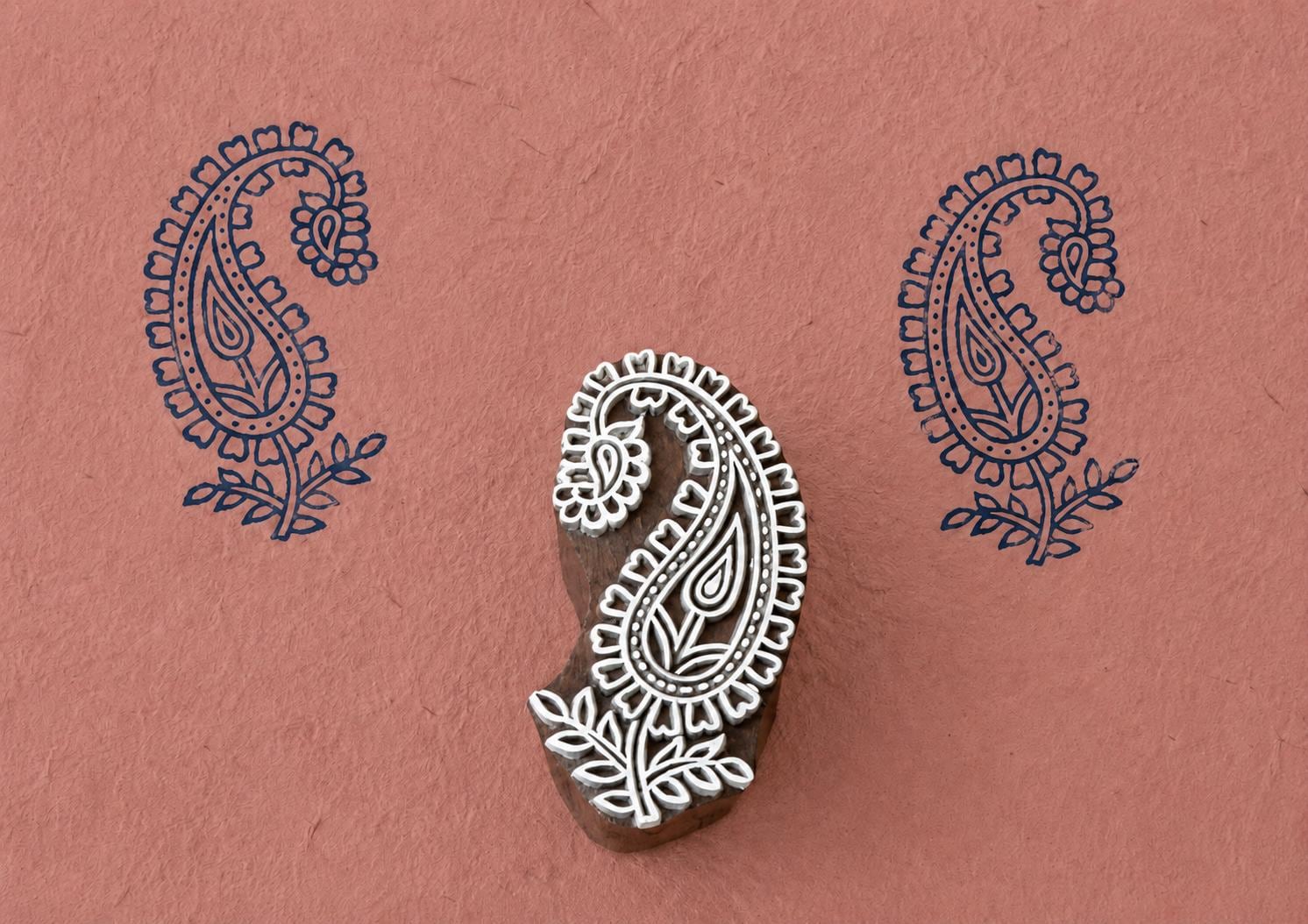 Hand-carved wooden printing block featuring a paisley flower motif with leaves, stamped in navy blue on textured terracotta handmade paper.