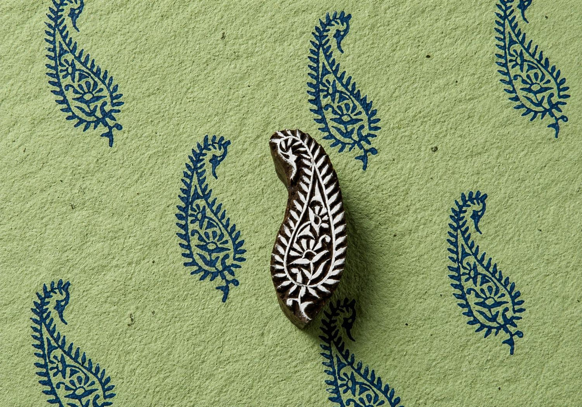Hand carved wooden paisley printing block displayed with repeating blue floral paisley impressions on textured handmade paper.