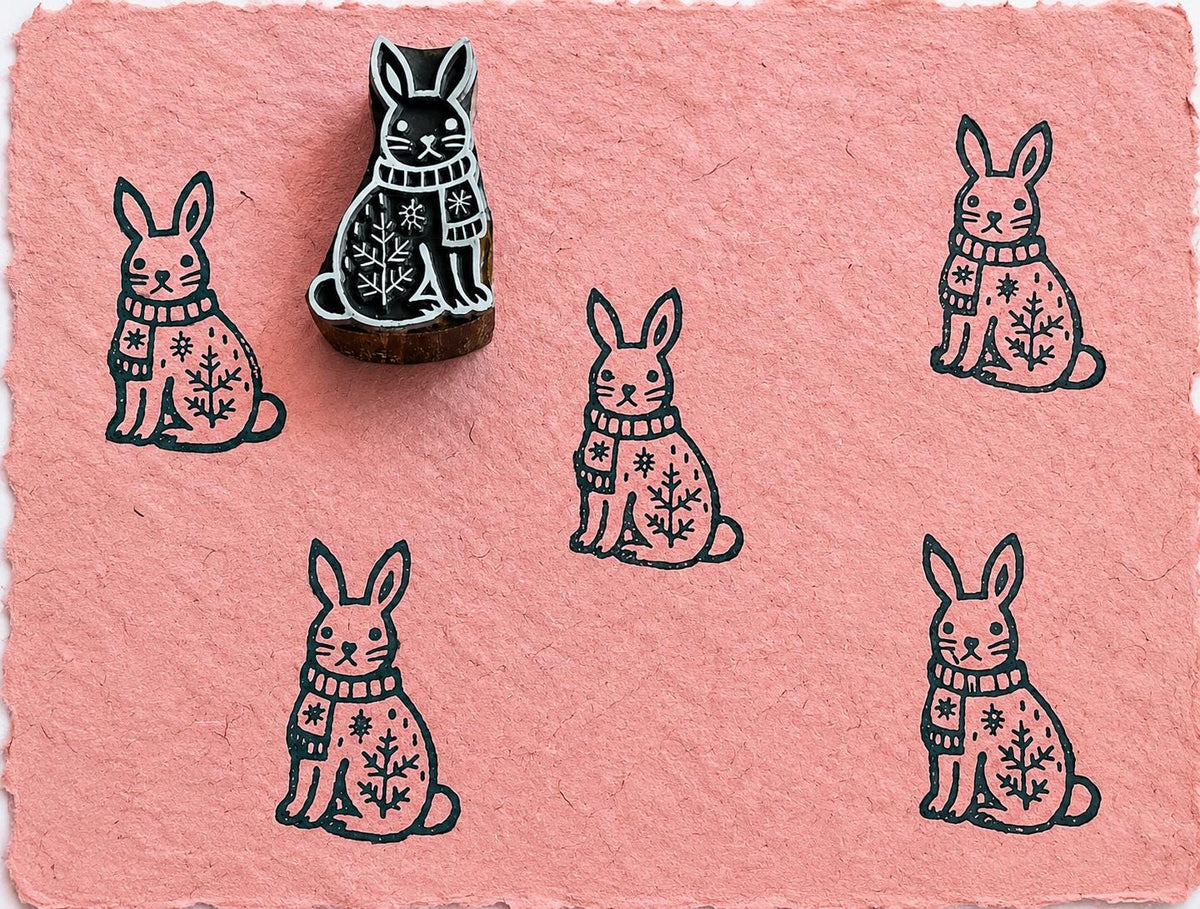 Hand carved rabbit printing block shown with repeating rabbit impressions on textured pink handmade paper.