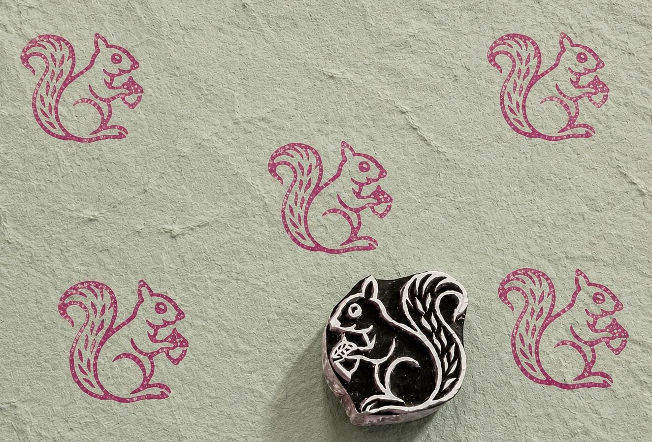 Hand-carved wooden printing block featuring a squirrel holding an acorn, stamped in berry pink ink on textured handmade paper.