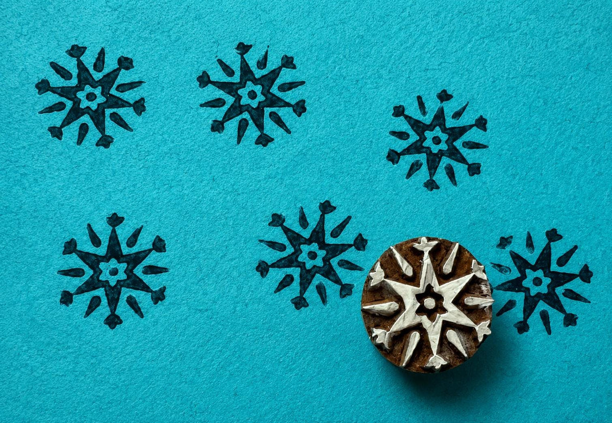 Hand carved Winter Star wooden block stamp creating repeated folk snowflake prints on textured blue handmade paper.