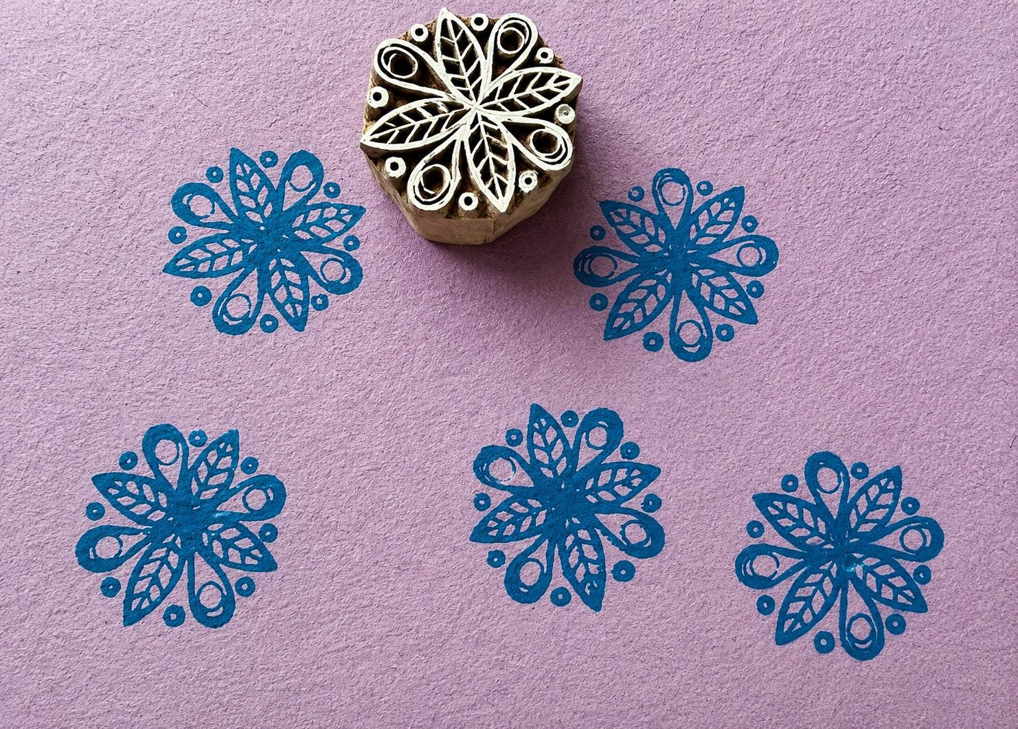 Hand carved wooden ornamental starburst printing block with blue geometric impressions on textured lavender handmade paper.