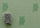 Hand-carved wooden printing block stamp featuring a folk owl design with detailed feathers and large circular eyes, shown stamped on textured sage green handmade paper.