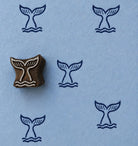 Hand carved whale tail wooden block stamp displayed on blue handmade paper with repeating ocean-inspired impressions.