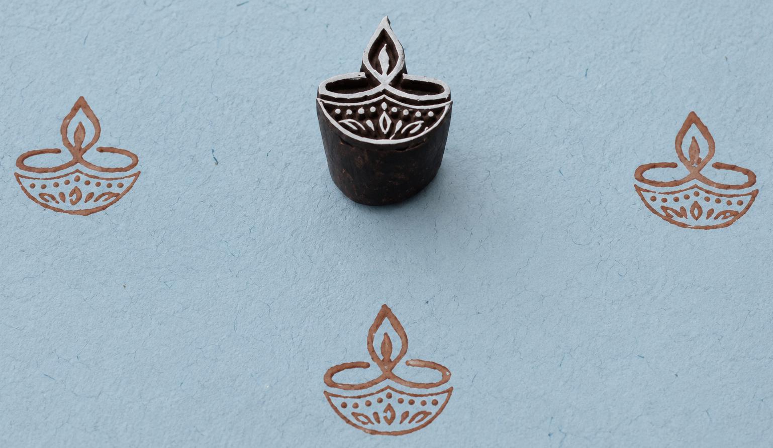 Hand-carved wooden printing block featuring a traditional diya lamp motif stamped in brown ink on light blue handmade paper.