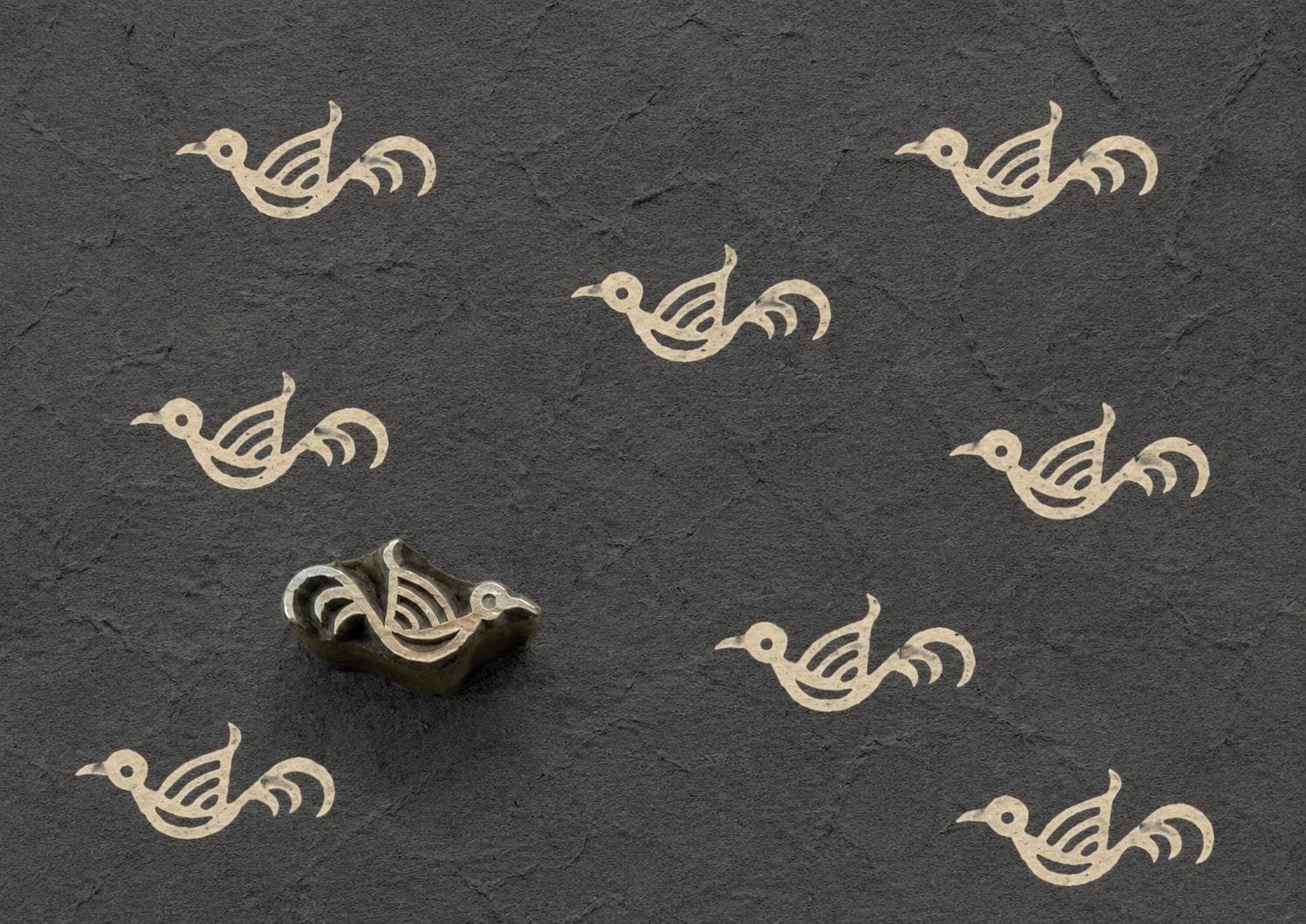 Hand-carved wooden printing block featuring a simple folk bird motif stamped repeatedly in ivory ink on textured charcoal handmade paper.