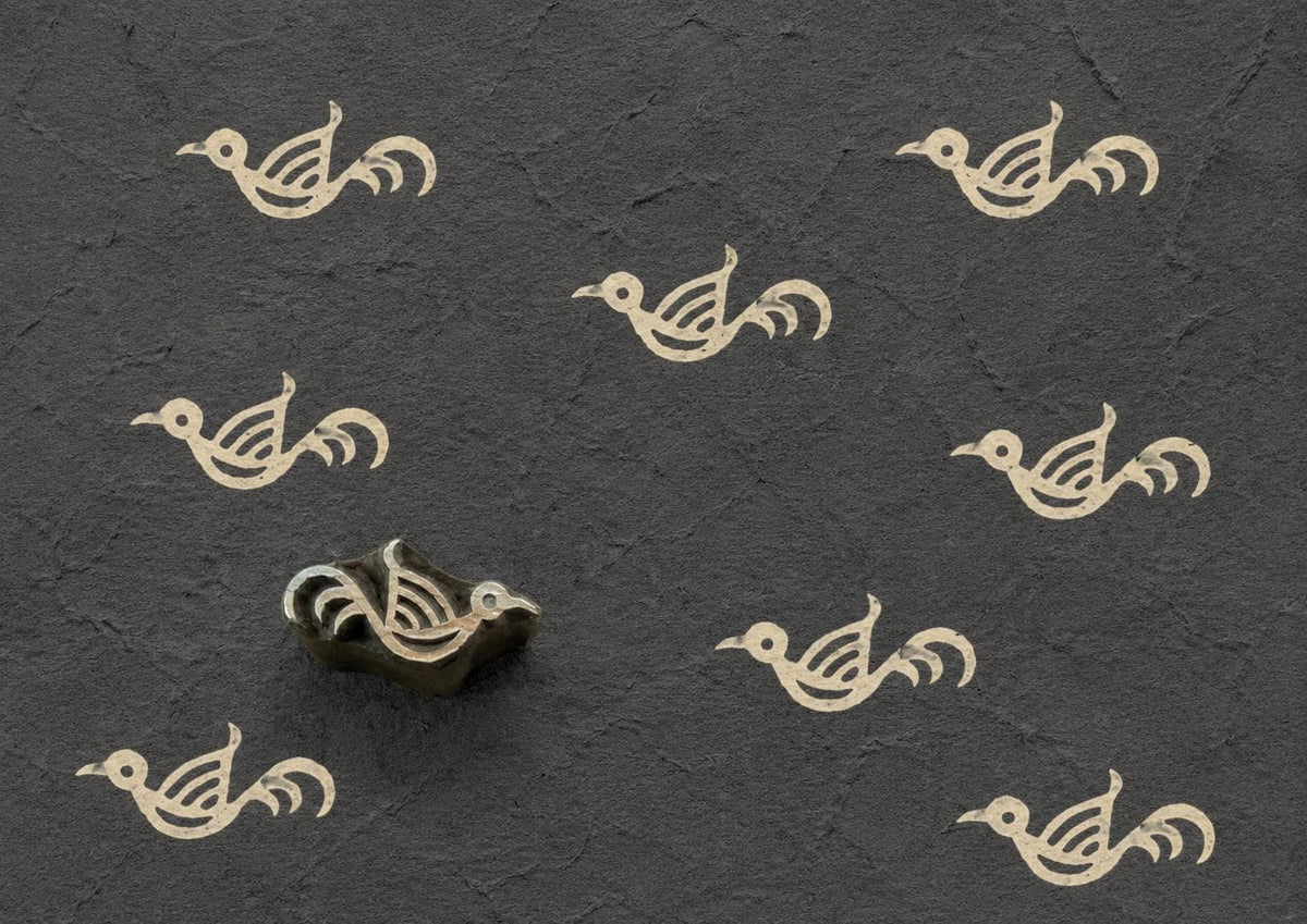 Hand-carved wooden printing block featuring a simple folk bird motif stamped repeatedly in ivory ink on textured charcoal handmade paper.