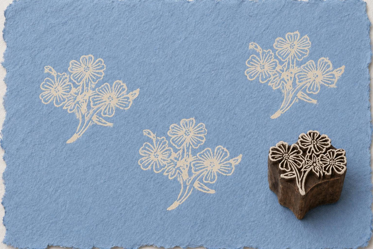 Hand-carved wooden printing block featuring a wildflower bouquet design stamped in ivory ink on textured blue handmade paper.