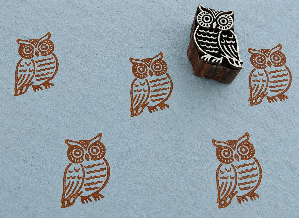 Hand-carved wooden printing block featuring a barn owl design with large decorative eyes and patterned feathers, stamped in brown ink on handmade blue paper.