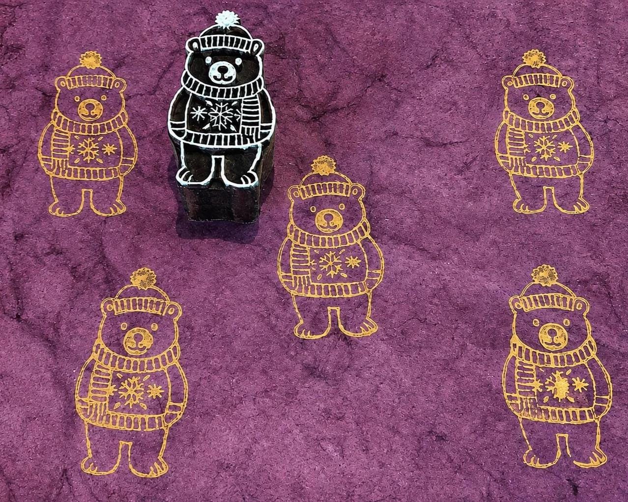 Hand-carved wooden winter bear printing block featuring a bear in a knitted sweater and hat, stamped in gold on purple handmade paper.