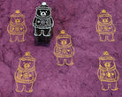 Hand-carved wooden winter bear printing block featuring a bear in a knitted sweater and hat, stamped in gold on purple handmade paper.