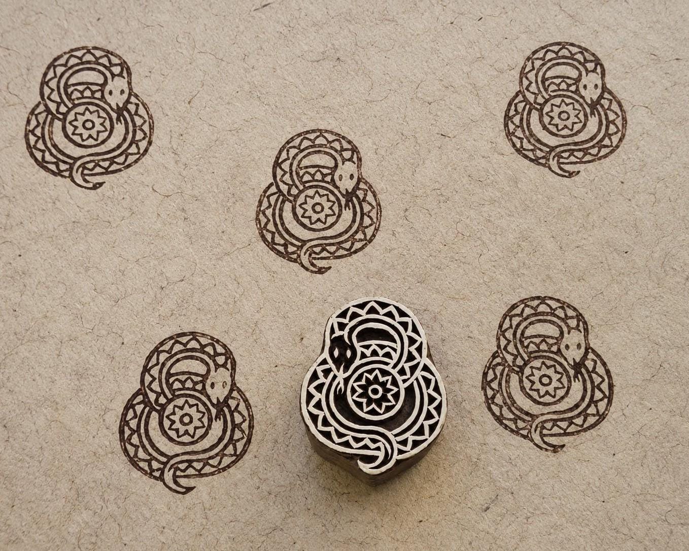 Hand-carved wooden folk art snake printing block with repeated brown coiled snake impressions on handmade paper.