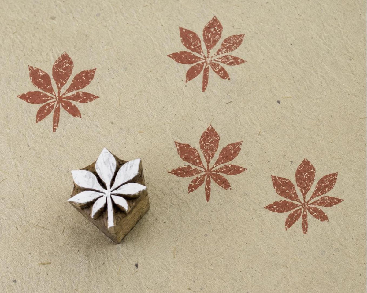 Hand carved wooden printing block featuring a seven petal bloom motif with terracotta stamped impressions on handmade paper.