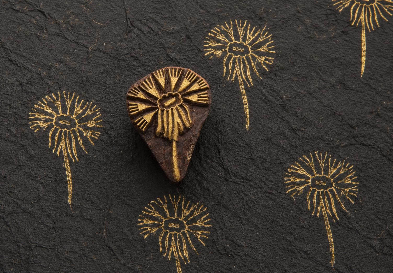 Hand carved wooden daisy bloom block printing stamp creating gold floral prints on handmade black paper.