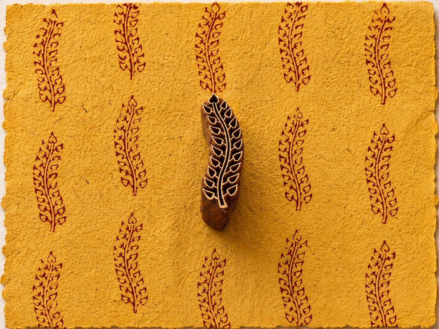 Hand carved wooden printing block featuring a paisley-inspired fern leaf design stamped repeatedly on mustard handmade paper using burgundy ink.