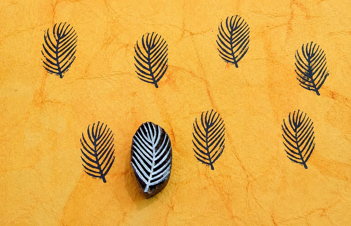 Hand-carved wooden tropical leaf printing block with repeated navy blue leaf impressions on handmade paper, traditional Indian block print stamp.