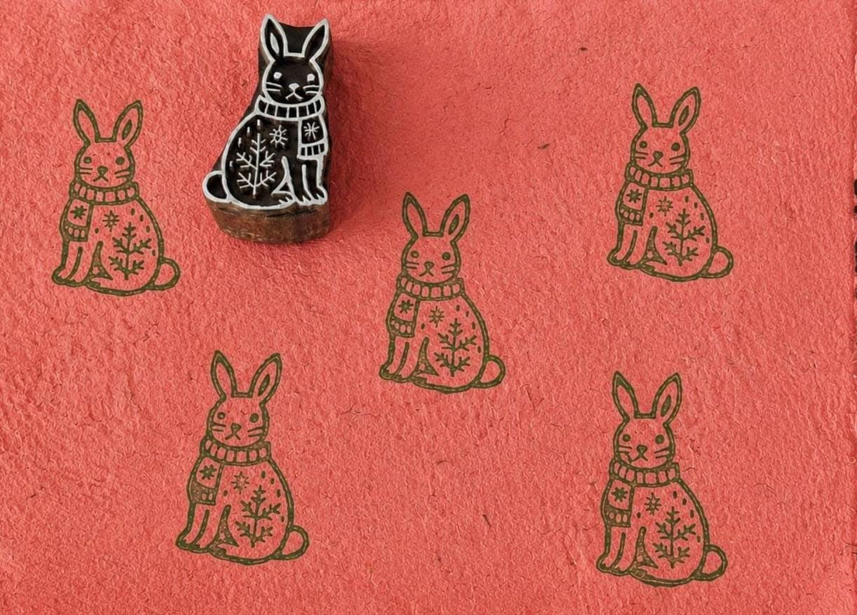 Hand-carved wooden winter bunny printing block with sweater and snowflake design, stamped repeatedly on coral handmade paper for Christmas fabric and paper crafts.