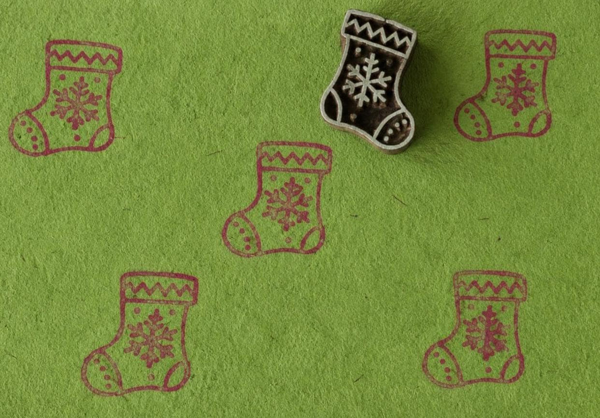 Hand-carved wooden Christmas stocking printing block with repeated festive stocking impressions on bright green handmade paper, ideal for holiday fabric printing and paper crafts.