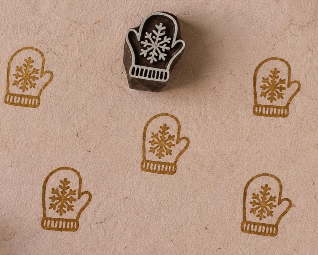 Hand-carved wooden snowflake mitten printing block with repeated mitten impressions on natural handmade paper, ideal for Christmas fabric printing, gift wrap, and holiday paper crafts.