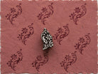 Hand-carved paisley floral vine wooden printing block surrounded by repeated dark burgundy floral impressions on textured dusty rose handmade paper.
