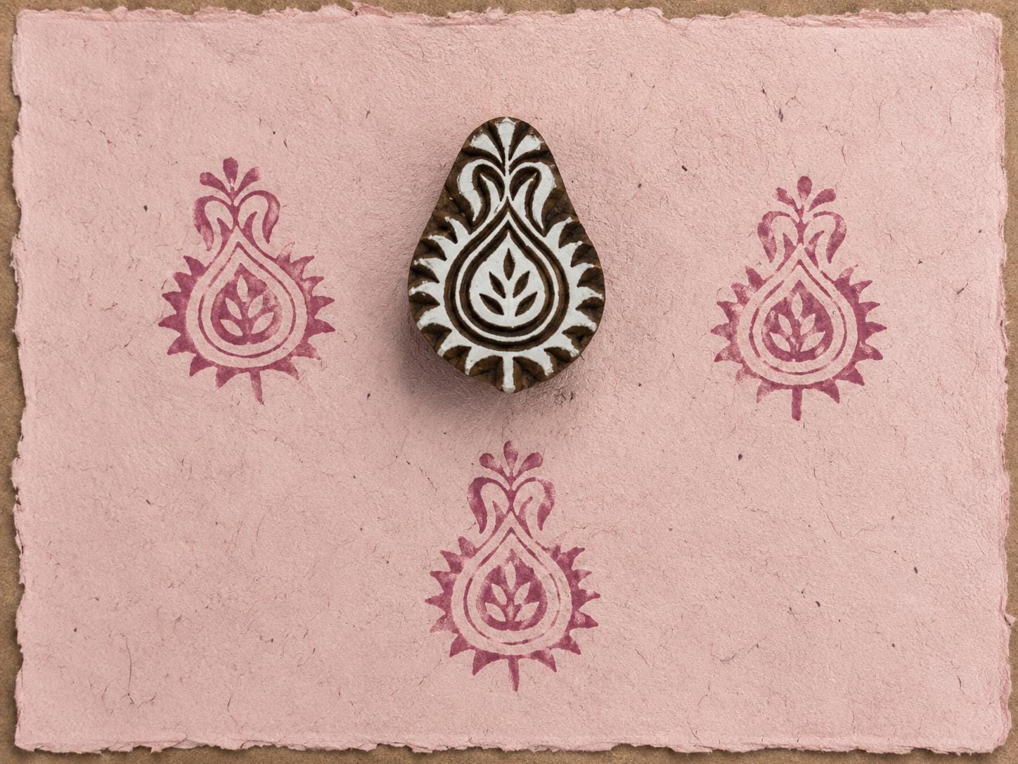 Hand-carved Indian paisley wooden printing block displayed on pink handmade paper with repeating berry-colored paisley impressions.