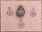 Hand-carved Indian paisley wooden printing block displayed on pink handmade paper with repeating berry-colored paisley impressions.