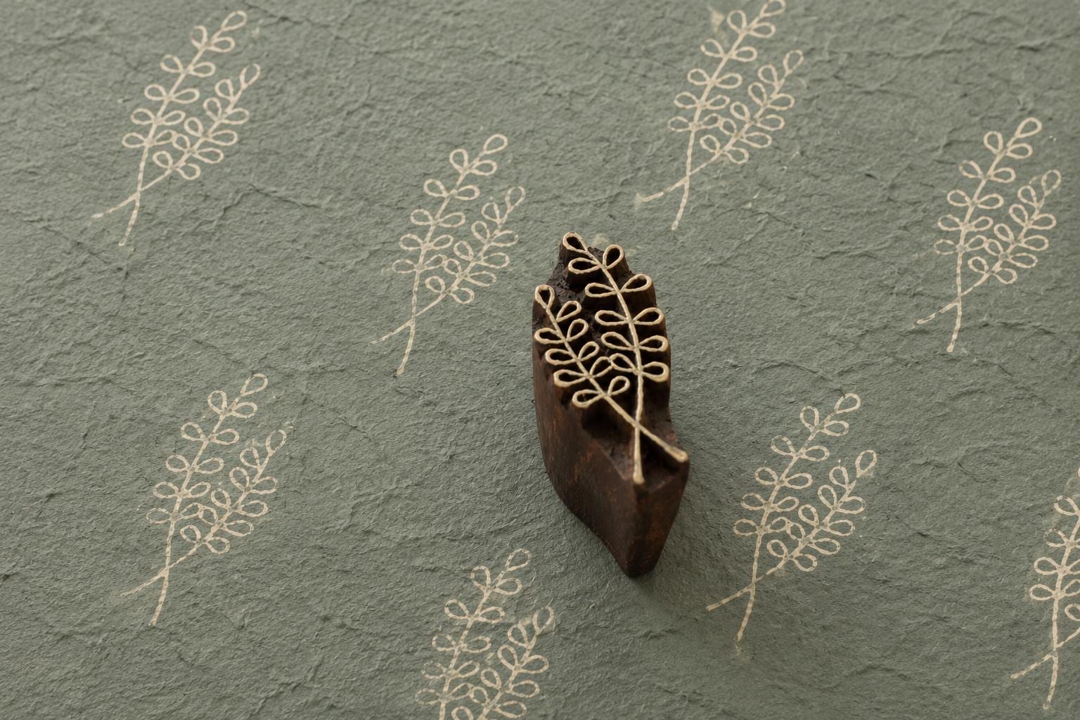 Hand carved wooden printing block featuring a delicate willow fern sprig botanical design stamped repeatedly on textured handmade sage paper.