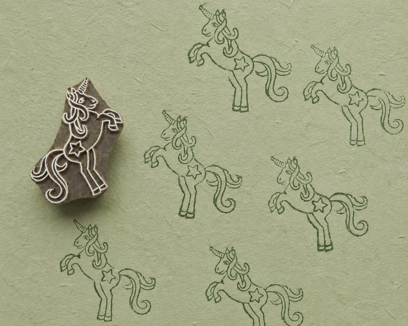 Hand carved wooden printing block featuring a prancing unicorn design with repeated dark green impressions on textured sage handmade paper.