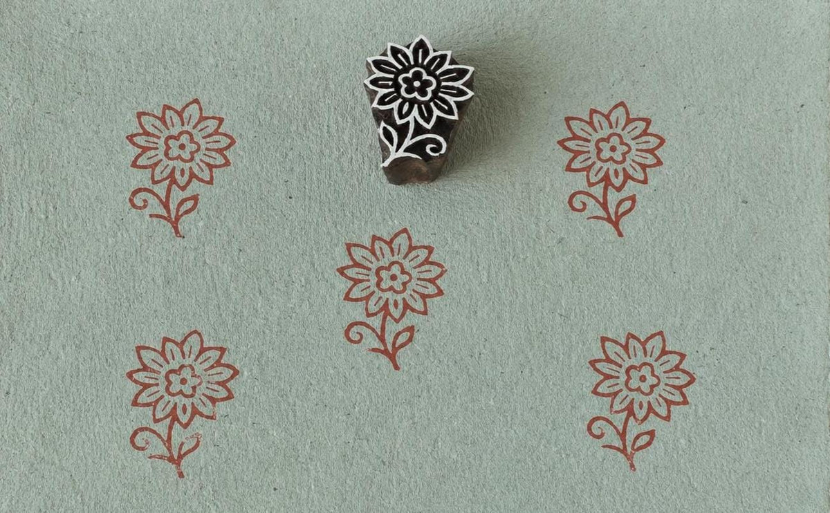 Hand-carved wooden floral printing block with repeated rustic blossom impressions on sage green handmade paper, ideal for fabric printing, stationery, and DIY crafts.